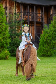 A little girl in the Ukrainian national costume rides a pony on the lawn