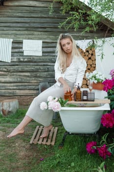 A woman is sitting on a cast-iron bathtub in the courtyard of a country house next to a bush of flowering peonies. The concept of summer, country life, a bathroom on the street in a blooming garden in the country