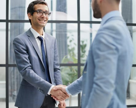 Businessmen shake hands, standing near a large office window