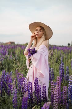 A beautiful woman in a straw hat walks in a field with purple flowers. A walk in nature in the lupin field.