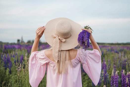 A beautiful woman in a straw hat walks in a field with purple flowers. A walk in nature in the lupin field.