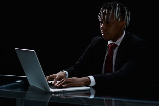 Focused dark-skinned businessman sits at a table in a modern office, uses a laptop, conducts business correspondence, answers emails