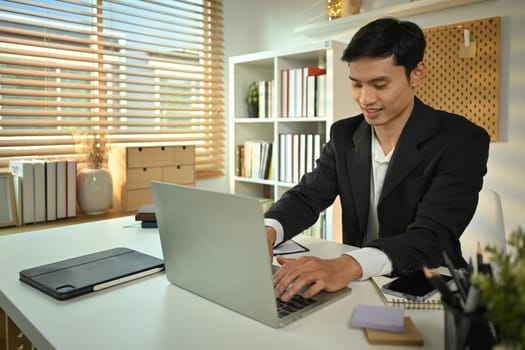 Handsome male manager in black suit typing on laptop, searching online information at workplace.