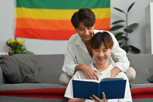 Happy moment of two young male lovers embracing each other and reading book in living room. Homosexual and love concept.