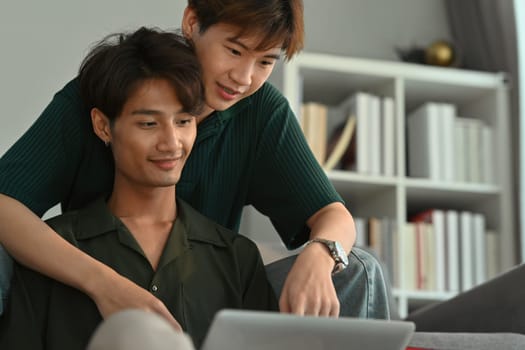 Happy same sex male couple embracing and using laptop in living room. LGBT, love and lifestyle relationship concept.