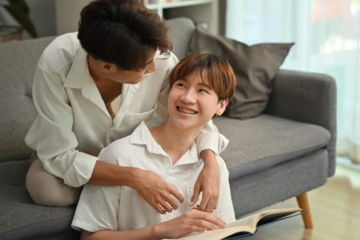 Peaceful two young male lovers embracing each other and reading book in cozy living room. Homosexual and love concept.