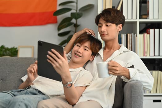 Loving homosexual couple watching videos or browsing internet on digital tablet while sitting on couch. LGBT, love and lifestyle concept.