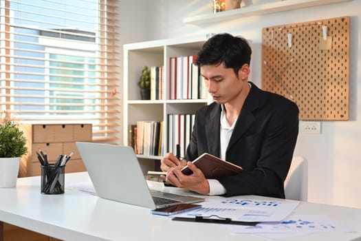 Focused male entrepreneur writing his strategy ideas on notebook and using laptop at working desk.