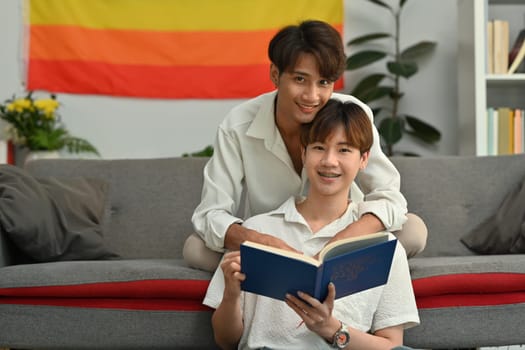 Peaceful two young male lovers embracing each other and reading book in cozy living room. Homosexual and love concept.