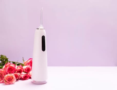 Dental discount, sale, holiday concept. Oral teeth irrigator, roses on purple background. Water tooth cleaner, white portable rechargeable cordless water dental flosser.Home dental care device. Vertical