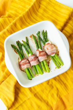 Asparagus baked with bacon and various spices. A bright background, an orange napkin on which the dish stands. Vertical photo