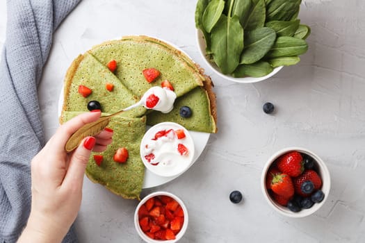 A woman's hand adds sour cream with berries to green spinach pancakes that lie on a plate with fresh berries and sour cream, on a gray background with textiles. copy space