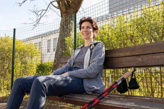 Smiling with Teeth Woman Sits on Bench Holding Poles After Nordic Walking. Beautiful Happy Caucasian Woman Enjoys Sunny Day. Physical Activity With Walking Poles. Outdoor Recreation. Horizontal Plane