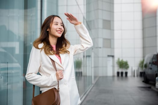 Portrait of confidence young businesswoman standing outside office building in city raise her hand to shade sun UV. Happy woman wearing white suit jacket with brown bag at sunlight outdoors. sun block