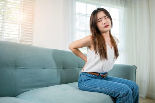 Asian woman holding her lower back feeling painful waist hurt massage in living room, beautiful young female touching back pain sitting in sofa she suffering from backache at home, healthcare medical