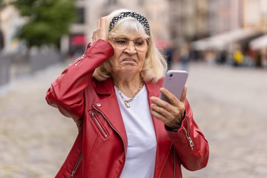 Senior woman use smartphone typing browsing, loses becoming surprised sudden lottery results, bad news, fortune loss, fail. Elderly grandmother walking in urban city street outdoors. Town lifestyles