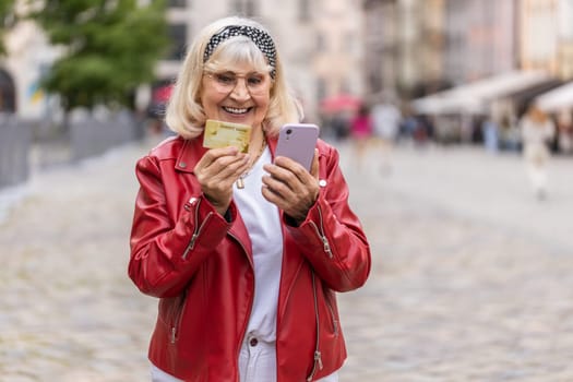 Senior woman using credit bank card smartphone while transferring money, purchases online shopping, order food delivery, booking hotel room. Elderly grandmother walking in urban city street outdoors
