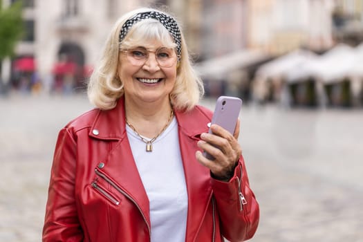 Smiling senior woman using smartphone typing text messages browsing internet, finishing work, looking at camera. Elderly grandmother walking in urban city street background. Town outdoors. Copy-space