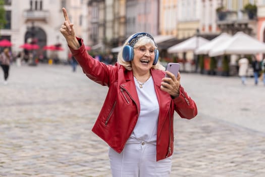 Happy overjoyed senior woman in wireless headphones listening favorite energetic music, celebrating win. Elderly grandmother tourist traveler walking in urban city street. Town lifestyles outdoors