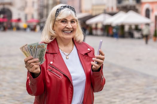 Happy senior woman counting money dollar cash, use smartphone calculator app, satisfied of income for planned vacation gifts. Elderly grandmother walking in urban city street. Town lifestyles outdoors