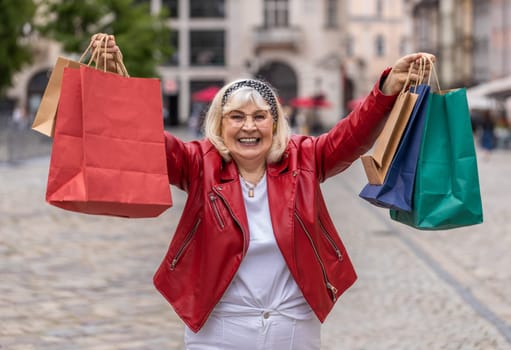 Happy smiling stylish senior woman shopaholic consumer after shopping sale with full bags with gifts outdoors. Elderly grandmother tourist traveler walking in urban city street road. Town lifestyles
