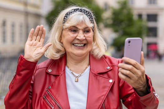 Senior woman blogger taking selfie on smartphone, communicating video call online with subscribers, recording stories for social media vlog outdoors. Elderly grandmother walking in urban city street