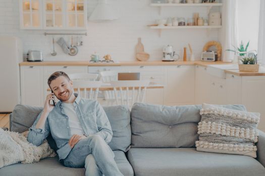 Happy smiling young man with stubble in casual clothes talking on phone with his friend while sitting with crossed legs on cozy couch, kitchen counter in blurred background. Communication concept