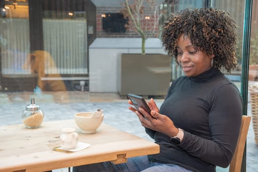 young african american woman sitting in a cafe with a cup of hot coffee, watching a social media feed in a smartphone, using technology, high quality photo, High quality photo