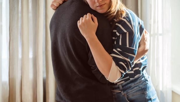 A young girl warmly and tenderly hugs her boyfriend, a couple stands against the background of a window on a sunny day, a woman begins romantic relationship. Husband and wife feel united, in harmony.