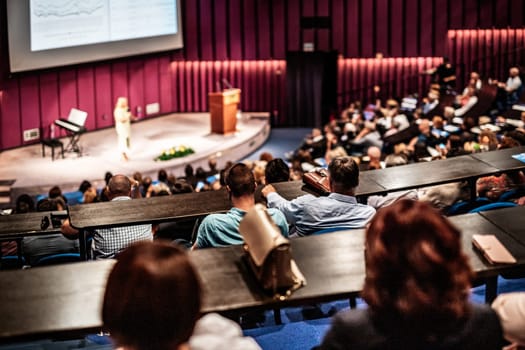 Business and entrepreneurship symposium. Female speaker giving a talk at business meeting. Audience in conference hall. Rear view of unrecognized participant in audience.