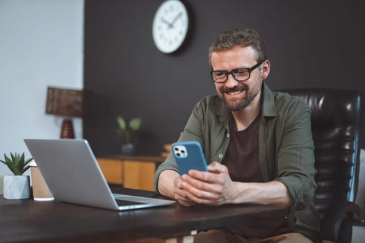 Smiling and happy mid-aged man engages in online activities while sitting at home on desk near laptop. Man is depicted multitasking, either sending text message or spending time online through phone. . High quality photo