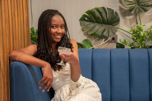 young afro woman sitting in a restaurant armchair toasting with a cocktail and smiling at the camera very happy. High quality photo