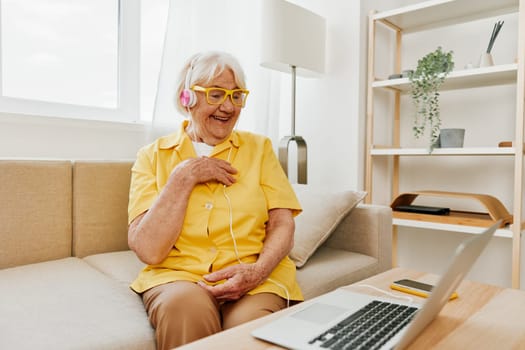 Elderly woman wearing headphones with a laptop communication online video call smile happiness, sitting on the couch at home and working in a yellow shirt, the lifestyle of a retired woman. High quality photo