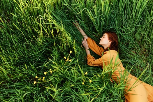 a relaxed woman enjoys summer lying in the tall green grass in a long orange dress stretched out stretching her arm forward. High quality photo