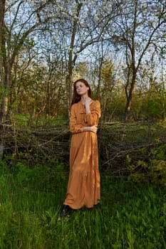 an elegant, sophisticated woman poses relaxed standing near a wicker fence at the dacha in a long orange dress, looking pleasantly at the camera. High quality photo