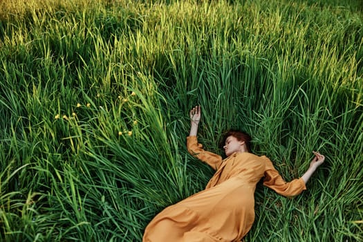 a sweet, calm woman in an orange dress lies in a green field with her arms outstretched, enjoying the silence and peace. Horizontal photo taken from above. High quality photo