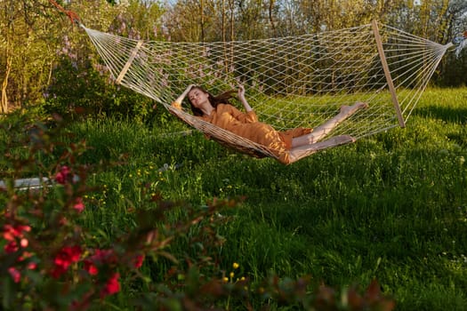 a happy woman in a long orange dress is relaxing in nature lying in a mesh hammock enjoying summer and vacation, looking at the sky. High quality photo