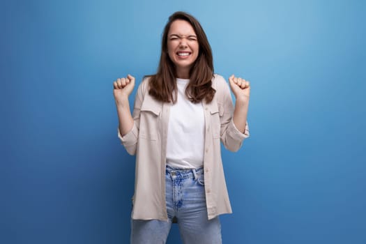 joyful happy young dark-haired lady in informal clothes celebrating victory on blue background.