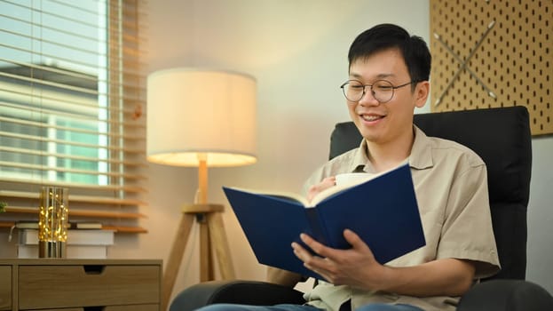 Peaceful millennial man in glasses sitting in comfortable armchair and reading book. Leisure and people concept.