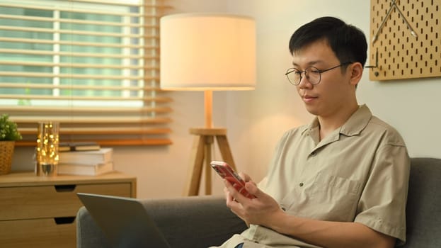 Image of man freelancer reading messages on mobile phone while sitting on couch with laptop in living room.