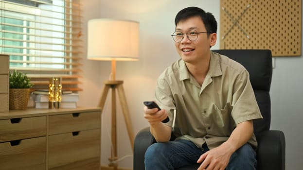 Smiling man in glasses and casual clothes holding remote control, watching TV in cozy living room.
