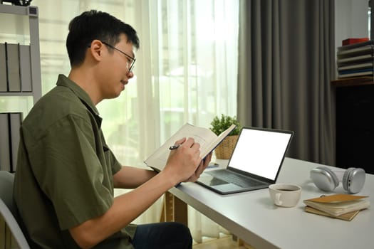 Male freelancer working remotely on laptop in living room, and making notes on notebook.