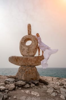 Woman sky stone. Lady stands on stone sculpture with ocean view. She is dressed in a white long dress, against the backdrop of the sea and sky. The dress develops in the wind