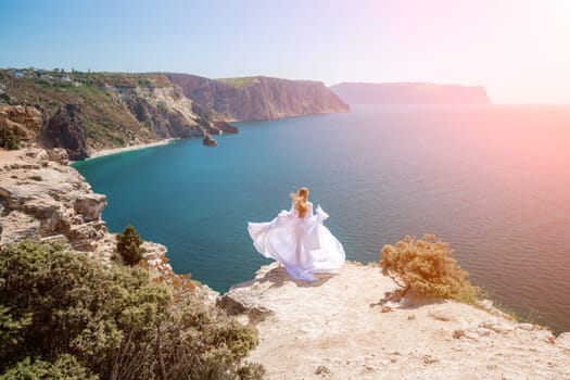 woman sea white dress. Blonde with long hair on a sunny seashore in a white flowing dress, rear view, silk fabric waving in the wind. Against the backdrop of the blue sky and mountains on the seashore