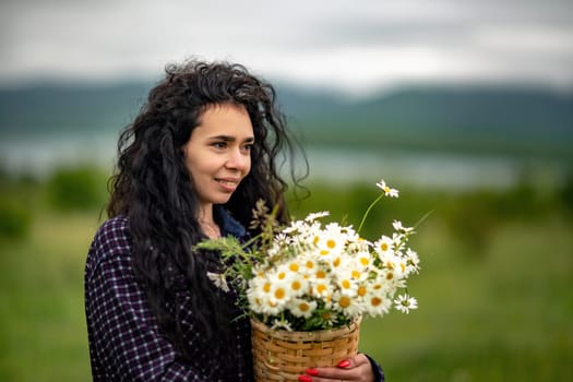 A woman stands on a green field and holds a basket with a large bouquet of daisies in her hands. In the background are mountains and a lake