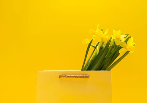 Bouquet of yellow narcissus in golden package, bag on yellow orange background. Spring sale, discount. International Women's Day celebration, Copy space for text. Monochrome minimalism. Horizontal