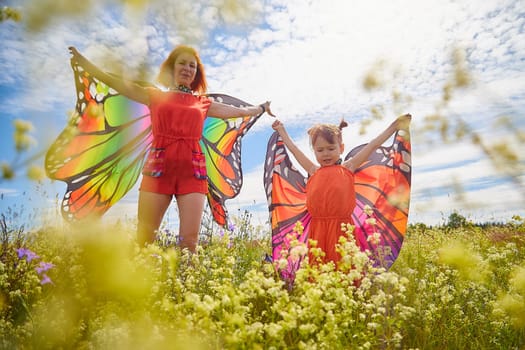 Happy female family with red haired mother and daughter with bright butterfly wings having fun on green and yellow meadow full of grass and flowers in sunny summer day. Concept family love