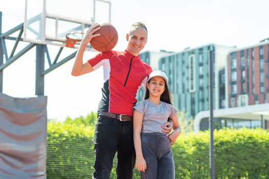 Happy father and teen daughter embracing and looking at camera outside at basketball court