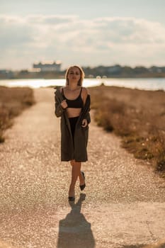 Portrait blonde sea cape. A calm young blonde in an unbuttoned khaki raincoat walks along the seashore, under a raincoat a black skirt and top.