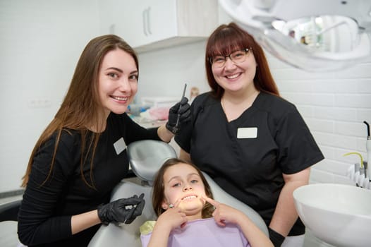 Female dentist hygienist and dental assistant smiling at camera, while using dental tools, examining teeth and oral cavity of a cute child showing her beautiful smile, sitting in the dentist's chair
